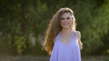 Güzel ve yetişkin bir kız kamera önünde aptallar, uzun kıvırcık saçlı bir Bayan dokunur, o bir sarafan giyinmiş ve doğada