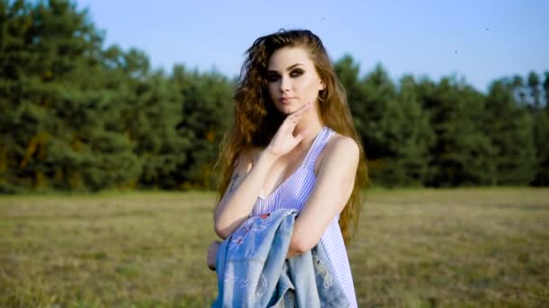 Uma jovem mulher em um sarafan de verão pensivamente fica na natureza em um dia de verão, uma senhora veste uma jaqueta de ganga em seu ombro — Vídeo de Stock