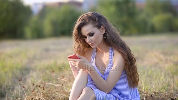 En romantisk kvinna med sotade ögon och lockigt hår skriver ett meddelande på sin mobiltelefon, en dam sitter på gräset i parken och skriver ut text på pekskärmen — Stockvideo