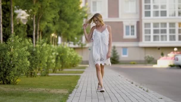 在炎热的夏天，将设计器穿白裙子的女人慢慢地走在街上，一位金发女郎走在人行道上 — 图库视频影像