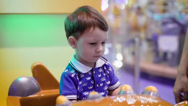 Маленький мальчик едет на большой игрушечной машине в парке аттракционов возле яркой круглой карусели, принимая реальные ключи от машины — стоковое видео