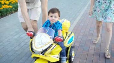 küçük çocuk parlak oyuncak motosiklet üzerinde bir şehir sokakta yaz günü ailesi tarafından eşlik kullanıyormuş