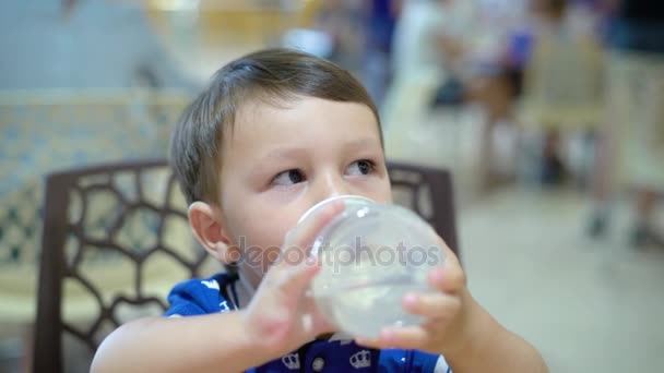 Λίγο σκοτεινός-eyed αγόρι κάθεται σε μια καρέκλα και το πόσιμο νερό από το μπιμπερό στο café, εσωτερικη — Αρχείο Βίντεο