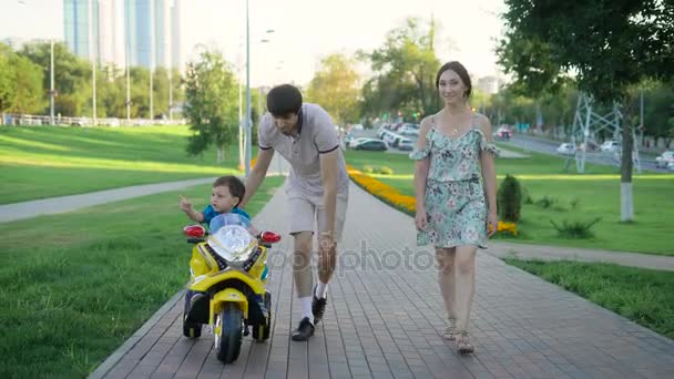 Familie spaziert an einem sonnigen Sommertag durch eine Gasse in der modernen Stadt, Vater hilft seinem kleinen Sohn beim Motorradfahren — Stockvideo