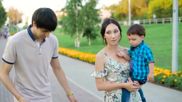 Отец, мать и их маленький сын гуляют в городском парке летом, разговаривают, улыбаются и смотрят друг на друга — стоковое видео