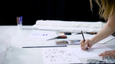 Genç sarışın kadın ayakta stüdyo ve çizim gelecekteki elbiseler terzilik içinde çalışma tablosu üzerinde bükme ile kapa. Giysi Tasarımcısı ile kalem çizimler yapıyor yeni moda koleksiyonu oluşturma.