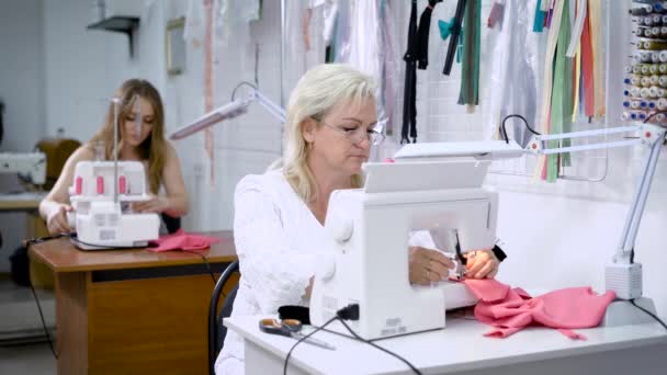 Κατασκευή δύο γυναίκες κάθονται στα τραπέζια εργασίας στα ρούχα. Γυναίκα ράψιμο ροζ ύφασμα στη σύγχρονη μηχανή και στοιχείο κοπής με ψαλίδι. Κορίτσι στο παρασκήνιο threading μια βελόνα για τη χρήση overlock. — Αρχείο Βίντεο