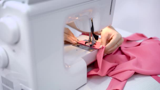Primer plano de las manos de la costurera que trabaja con la máquina de coser en el estudio de sastrería. Proceso de costura de tela rosa. Los dedos femeninos sostienen el material y hacen líneas con aguja e hilo . — Vídeos de Stock