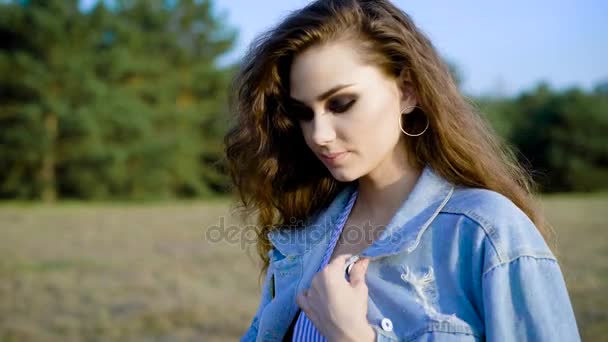 Молодая очаровательная женщина отправляется на довольно долгую прогулку по полю в джинсовой куртке и красиво улыбаясь солнцу — стоковое видео