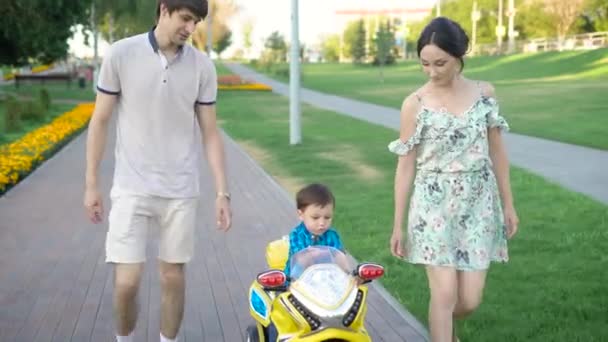 Feliz familia caminando con su hijo pequeño en el coche de juego en el parque. Niño disfrutando de jugar coche mientras monta con los padres en la pasarela en el parque . — Vídeos de Stock