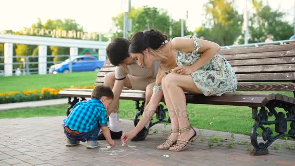 Giovani genitori in posa su panchina e disegno su asfalto con figlio. Giovane uomo e donna seduti sulla panchina e aiutare il piccolo figlio a disegnare con gesso a terra . — Video Stock