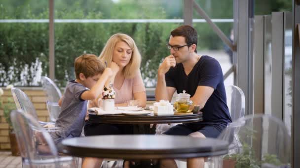 Szczęśliwa rodzina z syna, spędzać czas razem w odkrytym tarasem. Żona i mąż mówi podczas chłopiec jedzenia deser ze śmietaną i czekoladą. Młodzi rodzice cieszyć obiad z dzieckiem w restauracji. — Wideo stockowe