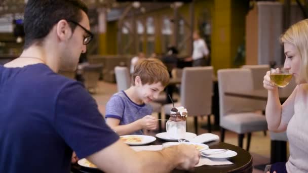 Una familia feliz con un hijo pequeño sentado a la mesa. Madre bebiendo té y usando su smartphone. Chico divirtiéndose comiendo postre de chocolate cremoso. Padres jóvenes disfrutando de la cena en restaurante con niño . — Vídeo de stock