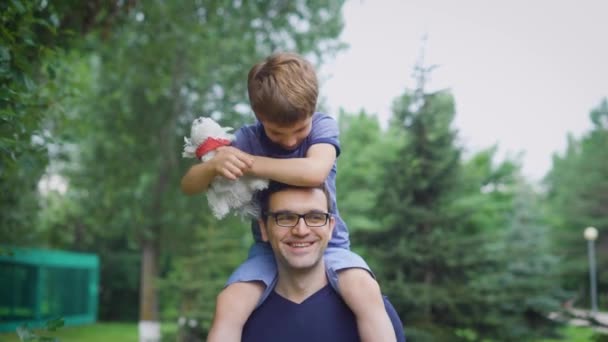 Knappe man in blauw shirt wandelen op straat met zijn zoon. Mannelijke bovenliggende tonen aan kind stedelijk landschap. Jongen zit op zijn vaders schouders bedrijf squishable hond en spelen. — Stockvideo