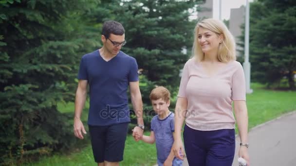 Primer plano de familia feliz caminando juntos en la calle riendo. Padre y madre hablando el uno con el otro y el hijo. Pareja cariñosa y su hijo pisando el camino con árboles en el fondo . — Vídeos de Stock