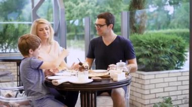 Küçük bir çocuk annesi bir sandviçle balık denemek için verir, kocası karısına aşık görünüyor, bir restoranda terasta aile akşam