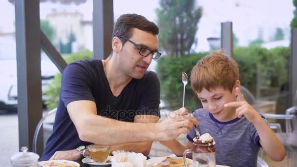 Un joven padre prueba sus hijos cóctel de leche con helado y chocolate, el hijo se ríe, la familia está cenando en el restaurante — Vídeo de stock
