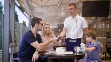 Genç yiyecek için ödemeli bir banka kartı ile eşleri, Garson terminal tutar, mutlu aile tastily restoranda yemek yedik, çocuk onun oyuncakla oynar