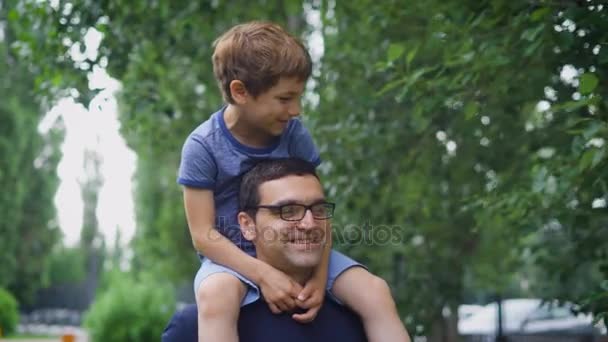 Un hombre feliz y adulto lleva a su pequeño hijo sobre sus hombros, a un niño le gusta pasar sus días de verano en una compañía masculina con su padre — Vídeos de Stock