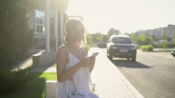 Стильна молода дівчина сидить на вулиці і переглядає смартфон, позує на сонячному світлі . — стокове відео