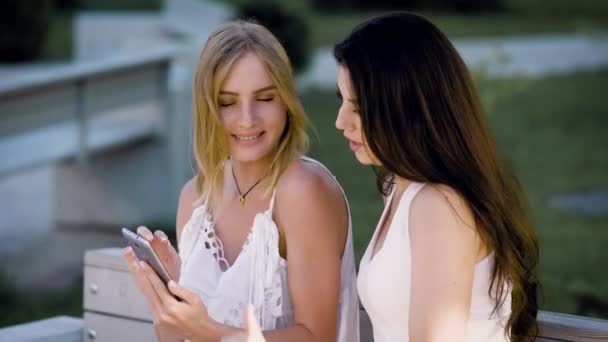 Dos mujeres jóvenes y elegantes sentadas en el banco y viendo el teléfono inteligente en verano . — Vídeo de stock