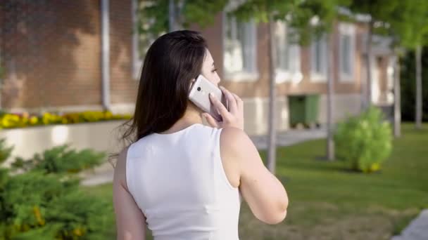 Vue arrière de brunette anonyme en blanc parlant smartphone sur fond de rue en été . — Video