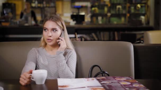Молодая и красивая женщина держит телефон в руке и общается с парнем, леди зашла в кафе выпить чашечку кофе — стоковое видео