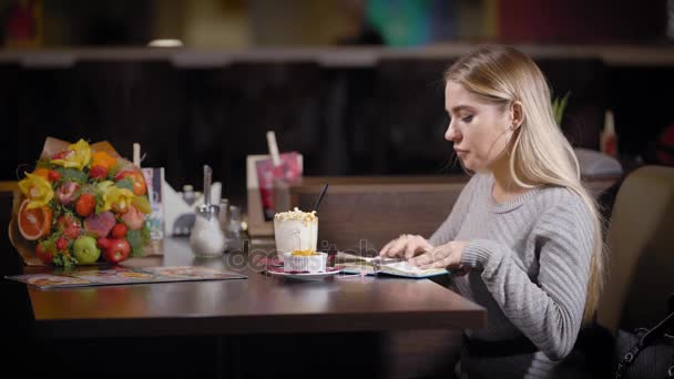 Een jonge en mooie vrouw is eten lunch in een restaurant, een dame voor een dessert met milkshakes, eten dat ze schreef op haar gedachten — Stockvideo