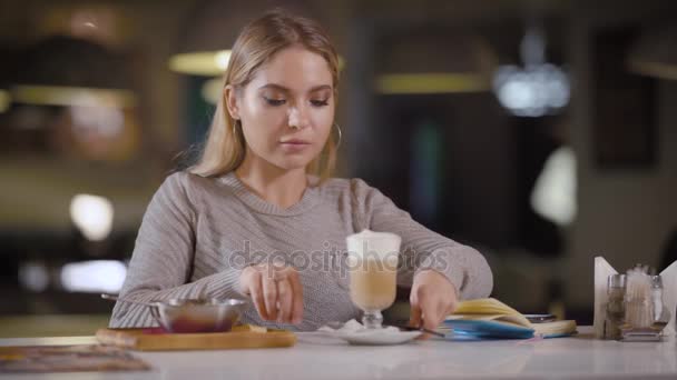 Genç ve güzel kadın bir restoranda lateks kahve içer, bir Bayan yiyor ve süt, bir içkiyle ona yalan bir not defteri sahiptir. — Stok video
