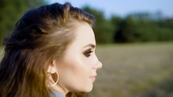 Vista lateral de la joven morena con maquillaje pesado con pendientes y mirando hacia otro lado en el fondo de la naturaleza . — Vídeo de stock