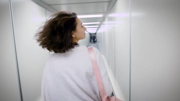 A woman with short and curly hair walks along a narrow corridor for boarding an aircraft, on her back is a stylish briefcase, she straightens her hair — Stock Video