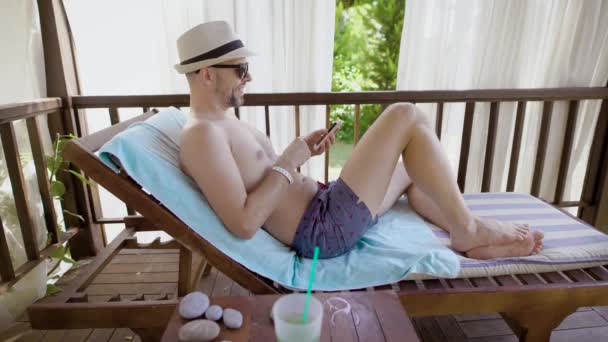 メガネで大人の男は、彼は日中は、彼の携帯電話で聞く音楽を楽しんで、人はリゾートでデッキチェアに横になっています。 — ストック動画