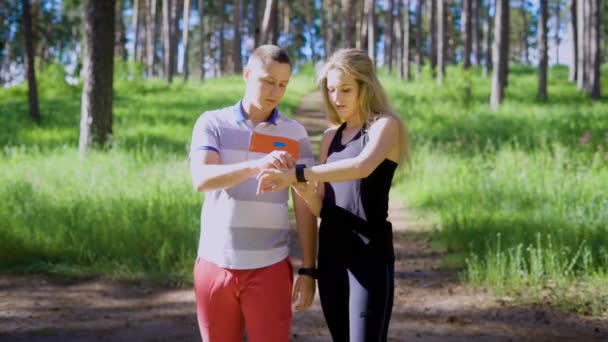 一对年轻夫妇在体育制服运行穿过森林在一个阳光明媚的日子，一个女运动员问男人什么距离他们克服了 — 图库视频影像