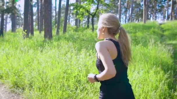 Slow motion shot of a young blonde woman in a sports suit, who actively runs through the park during the day, he enjoys the exercises — Stock Video