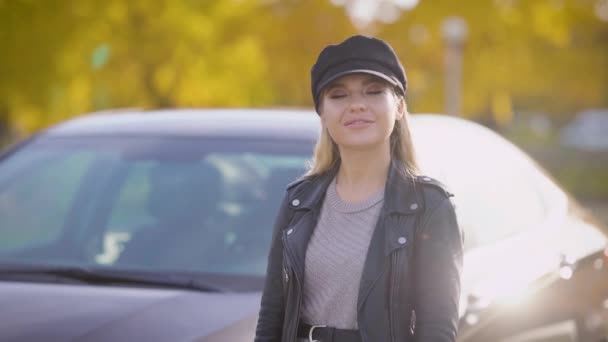 Stylish young woman with long straight blonde hair is wearing leather cap is standing on a fall street, smiling and looking at a camera — Stock Video