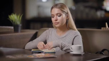 güzel mavi gözlü sarışın kız iç Cafe, alarak fincan kahve ve gülümseyerek, bir kameraya bakarak oturuyor