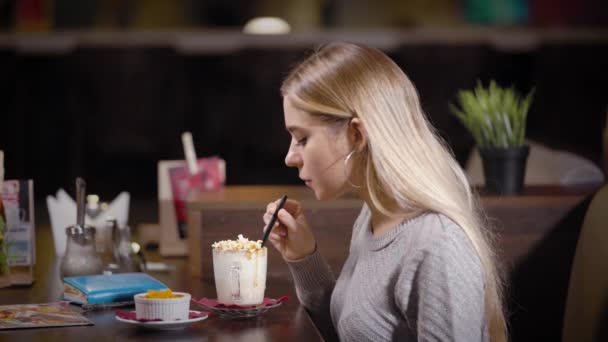 Charmante Blondine trinkt einen Kaffee mit Schlagsahne im Café, dann nimmt sie blauen Notizblock und Stift und schreibt Notizen — Stockvideo