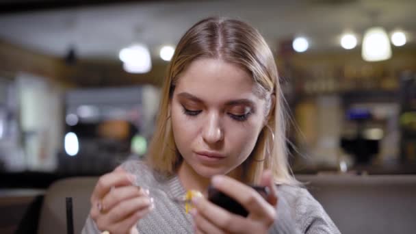 Mujer joven está comiendo postre en una mesa en la cafetería y viendo videos en la pantalla de su teléfono inteligente — Vídeos de Stock