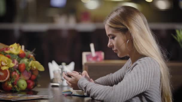 Ωραίο κορίτσι κάνει σημειώσεις στο σημειωματάριό της ενώ κάθεται σε ένα τραπέζι στο καφενείο, ψάχνει σε μια οθόνη των smartphone — Αρχείο Βίντεο
