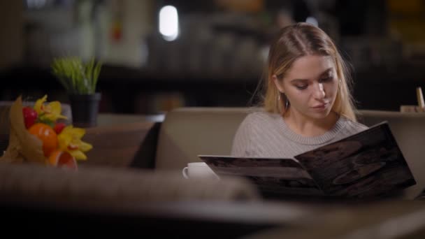 Einsame junge Frau mit blonden Haaren liest Speisekarte sorgfältig im Restaurant und wählt Mahlzeit zum Abendessen — Stockvideo