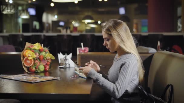 Zeker jonge vrouw poseren in café en schrijven in notebook — Stockvideo