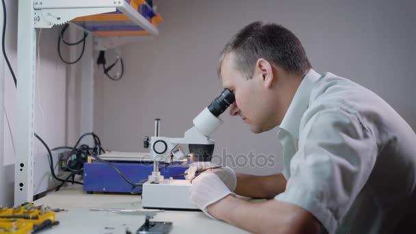 Electronic engineer is looking on a microscope in laboratory and studying small mechanism, touching details by tweezers — Stock Video