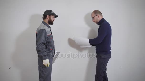 Cliente y reparador están de pie en un piso sobre un fondo de pared blanca, propietario está explicando qué hacer — Vídeo de stock