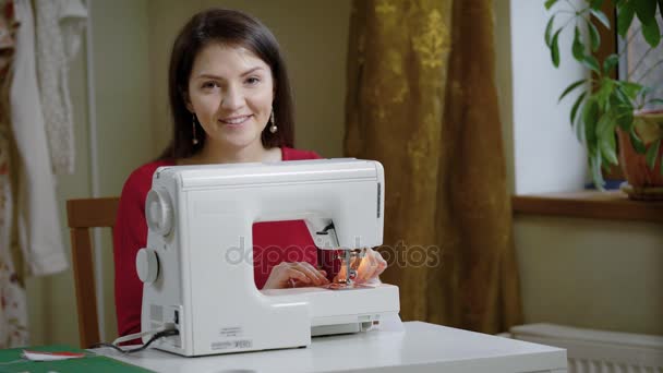 Glada vuxen kvinna med långt mörkt hår sitter vid ett bord med symaskin, tittar på en kamera — Stockvideo