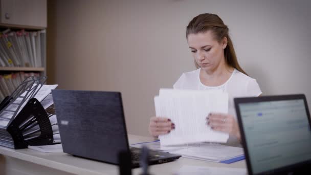 Uma jovem mulher que trabalha como contabilista em um escritório de advocacia examina documentos por trás de seu local de trabalho, antes de uma senhora fica um laptop — Vídeo de Stock