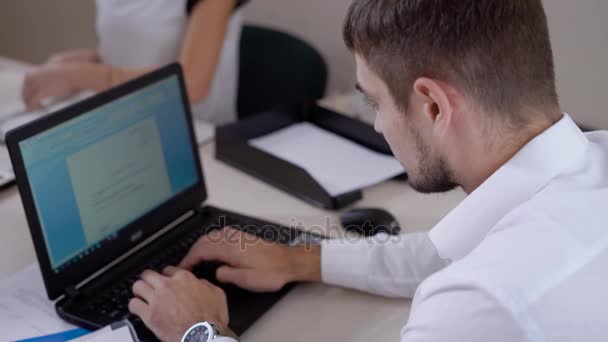 The young man prints the text on the laptop, the person looks at the laptop monitor and clicks on the keyboard buttons, he works in the office — Stock Video