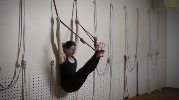 Beautiful brunette woman is hanging on the ropes leaning agains the wall in yoga centre. She is stretching her legs slowly going down. — Stock Video