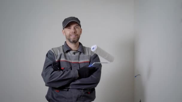 Веселий ремонтник стоїть в квартирі, тримаючи фарбувальний валик, перетинаючи руки на тлі білих порожніх стін — стокове відео