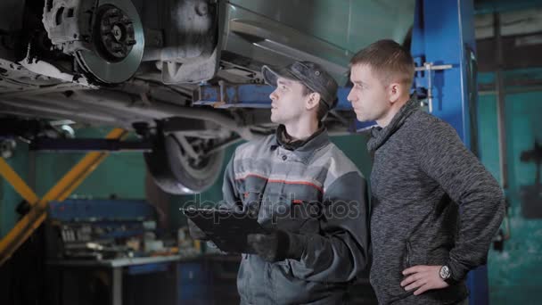 Bil mekaniker bedöma en bromsa disken, bromsbelägg och vattenslang, förklara för en bilägare och beräkna kostnaden för reparationen, i ett garage — Stockvideo