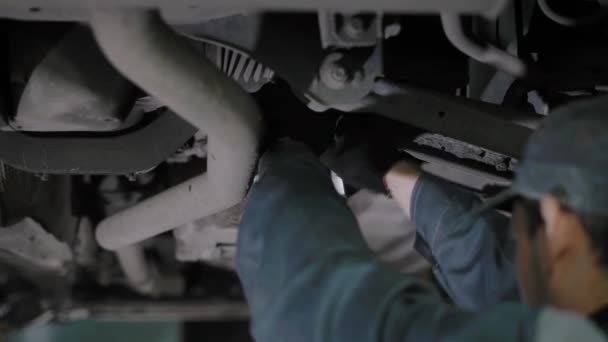 Mécanicien automobile règle le câble de frein à main, debout sous la voiture relevée, resserre les détails dans un atelier de réparation automobile — Video